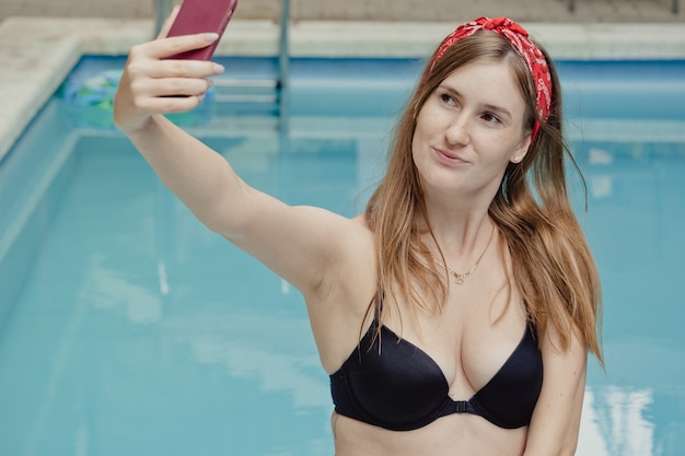 Mujer joven sonriente con teléfono en chaise. Relajante día de spa junto a la piscina en verano para hacer una foto o hacer una videollamada como novio