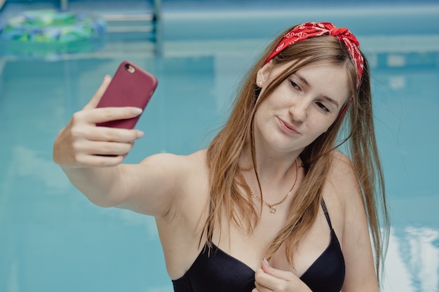 Mujer joven sonriente con teléfono en chaise. Relajante día de spa junto a la piscina en verano para hacer una foto o hacer una videollamada como novio