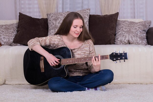 Mujer joven sonriente tocando la guitarra acústica negra en la alfombra en la habitación