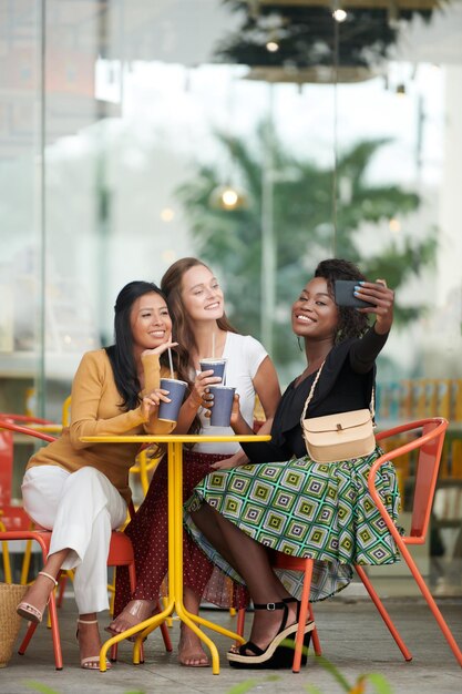 Mujer joven sonriente tomando selfie con sus mejores amigos cuando pasan tiempo en un café al aire libre