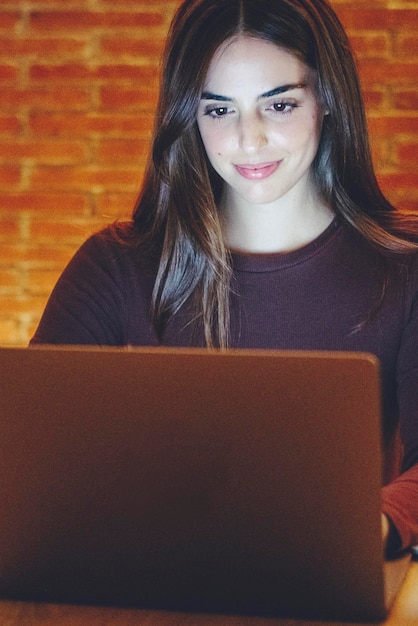 Mujer joven sonriente trabajando hasta tarde en la oficina de casa usando el espacio de copia de la computadora portátil Chica casual a la luz de la pantalla en el fondo de la habitación iluminada por luz amarilla Tecnología y concepto de trabajo