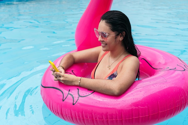 Mujer joven sonriente en traje de baño revisando su teléfono celular en una piscina en verano