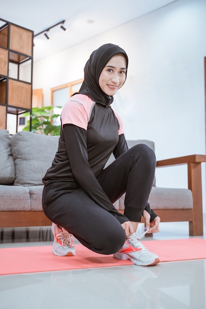 Mujer joven sonriente con un traje de entrenamiento hijab se pone en cuclillas mientras se coloca los cordones de los zapatos antes de hacer ejercicio en el interior