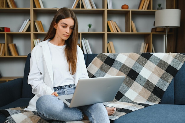 Mujer joven sonriente usando portátil sentada en el sofá en casa chica hermosa haciendo compras o chateando en línea en la red social divirtiéndose viendo películas freelance trabajando en un proyecto informático