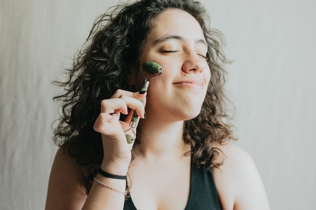 Mujer joven sonriente usando rodillo facial para el cuidado de la piel Hábitos de salud de belleza con masaje facial Preparándose y rutina diaria para una cara brillante Retrato de primer plano en el estudio