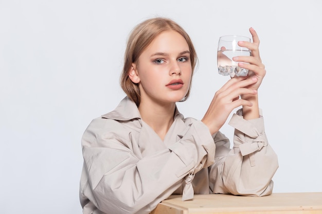 Mujer joven sonriente con vaso de agua hermosa chica en impermeable beige posando en estudio