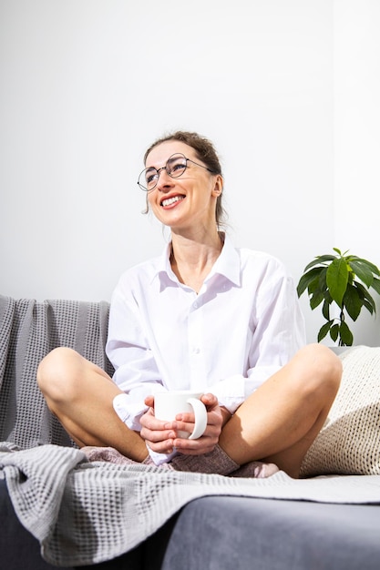 Mujer joven sonriente en vasos con una taza de café sentada en un sofá