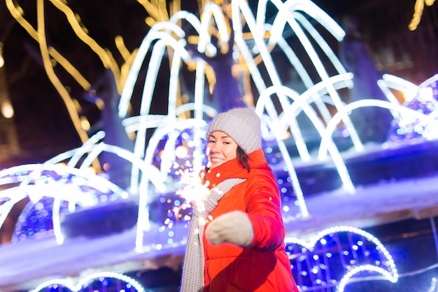 Mujer joven sonriente vistiendo ropa de punto de invierno sosteniendo bengala al aire libre sobre fondo de nieve. Vacaciones navideñas.