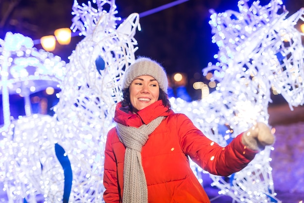 Mujer joven sonriente vistiendo ropa de punto de invierno sosteniendo bengala al aire libre sobre fondo de nieve. Vacaciones navideñas.