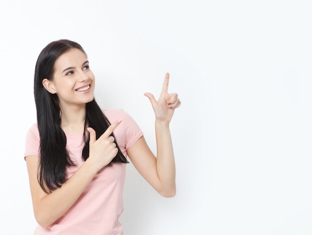 Mujer joven con sonrisa blanca perfecta en ropa de verano muestra los dedos índice a un lado en el espacio vacío sobre la pared blanca.