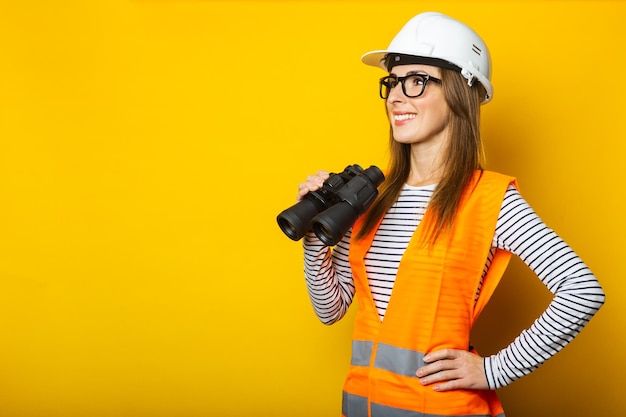 Mujer joven con una sonrisa en un chaleco y un casco sostiene binoculares sobre un fondo amarillo Concepto de construcción nuevo edificio Banner