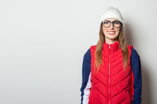 Mujer joven con una sonrisa en un chaleco rojo, sombrero y gafas contra una pared de luz