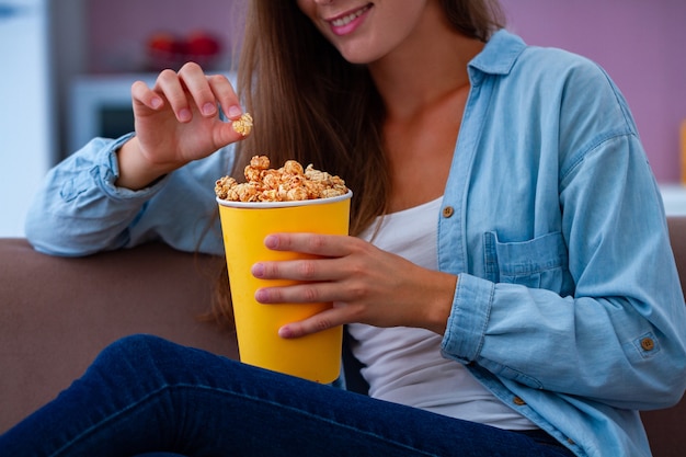 Mujer joven de la sonrisa feliz que descansa y que come las palomitas crujientes del caramelo durante mirar la TV en casa. Película de palomitas de maíz