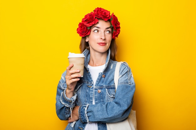 Mujer joven con una sonrisa mira hacia un lado con una chaqueta de mezclilla y una corona de flores rojas en la cabeza sostiene una taza de papel con café en una pared amarilla.
