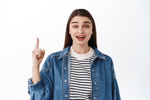 Mujer joven sorprendida apuntando con el dedo hacia arriba en el logo promocional superior, mostrando la pancarta hacia arriba y mirando al frente intrigada, emocionada con la promoción del evento, de pie sobre una pared blanca