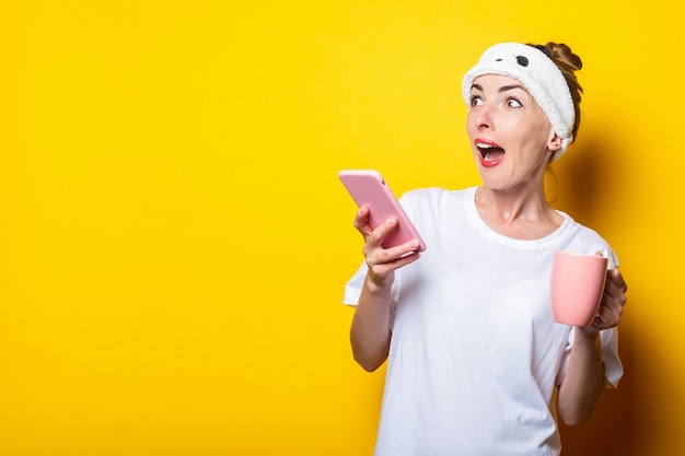 Mujer joven sorprendida asustada en un vendaje mira hacia el lado con un teléfono y una taza de café sobre un fondo amarillo