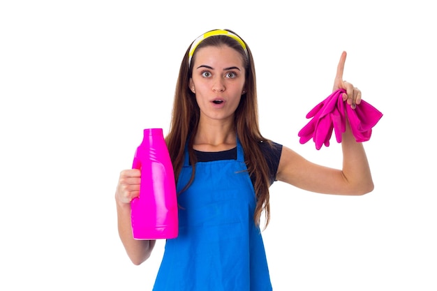 Mujer joven sorprendida en camiseta azul y delantal con guantes rosas y detergente en el estudio