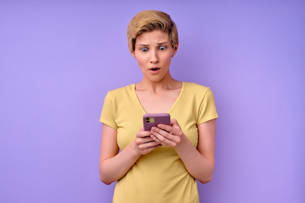 Mujer joven sorprendida en camiseta casual manteniendo la boca ampliamente abierta mirando al teléfono inteligente caucásico beau ...