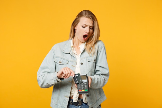 Mujer joven sorprendida y descontenta sostiene un terminal de pago bancario moderno inalámbrico para procesar, adquirir pagos con tarjeta de crédito aislados en un fondo naranja amarillo. Concepto de estilo de vida de las personas. Simulacros de espacio de copia.