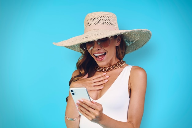 Mujer joven sorprendida en elegante sombrero mirando su teléfono móvil mientras está de pie contra el fondo azul