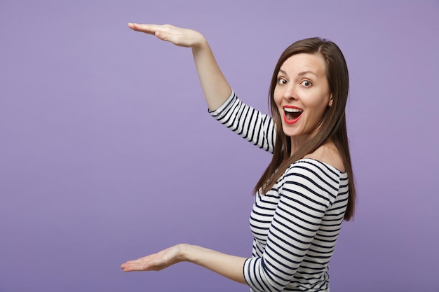 Mujer joven sorprendida y emocionada con ropa informal a rayas posando aislada en un fondo violeta violeta. Concepto de estilo de vida de las personas. Simulacros de espacio de copia. Gesticulando demostrando tamaño con espacio de trabajo vertical.