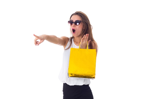 Mujer joven sorprendida en falda negra con gafas de sol sosteniendo bolsas de compras y señalando algo