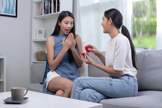 Mujer joven sorprendida levantando las manos y sonriendo mientras su novia le propone matrimonio en la sala de estar Pareja emocional enamorada compromiso Concepto de estilo de vida de pareja LGBT lesbiana asiática