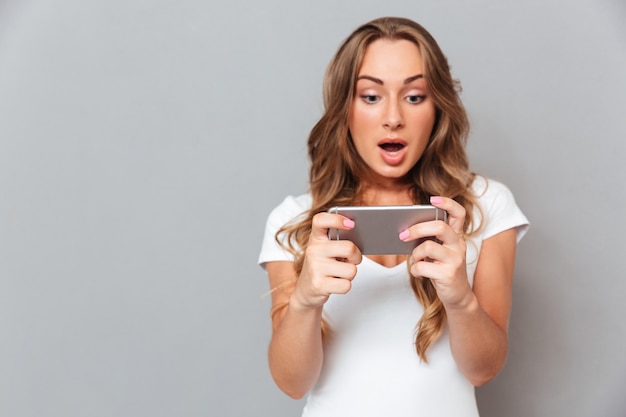 Mujer joven sorprendida mirando en smartphone aislado en una pared gris