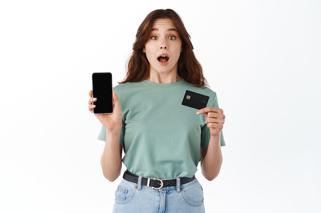 Mujer joven sorprendida que muestra la pantalla vacía del teléfono inteligente y una tarjeta de crédito plástica, demuestra la aplicación de banca móvil en línea, de pie contra la pared blanca