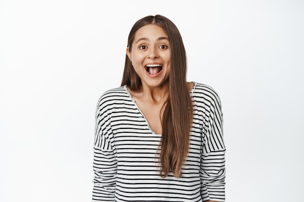 Mujer joven sorprendida que parece feliz y asombrada por la cámara, sonriendo emocionada, de pie sobre fondo blanco.