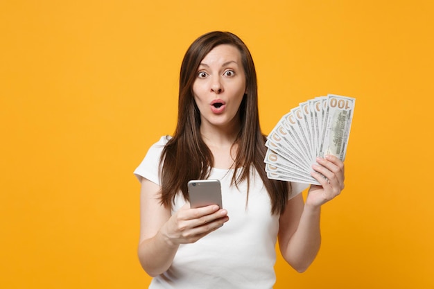 Mujer joven sorprendida con ropa blanca casual usando teléfono móvil, sosteniendo un ventilador de dinero en efectivo en billetes de dólar aislados en el fondo de la pared naranja amarilla. Concepto de estilo de vida de las personas. Simulacros de espacio de copia.