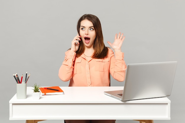 Mujer joven sorprendida con ropa casual pastel hablando por teléfono móvil sentada y trabajando en el escritorio con una computadora portátil aislada en un fondo gris. Concepto de estilo de vida de carrera empresarial de logro. Simulacros de espacio de copia.