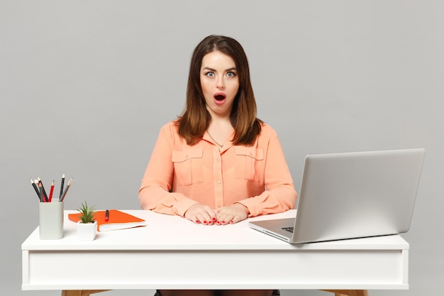 Mujer joven sorprendida con ropa casual pastel manteniendo la boca abierta sentada, trabajando en un escritorio blanco con una computadora portátil aislada en un fondo gris. Concepto de estilo de vida de carrera empresarial de logro. Simulacros de espacio de copia.