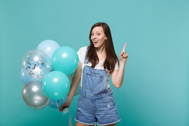 Mujer joven sorprendida con ropa de mezclilla apuntando con el dedo índice hacia arriba, celebrando y sosteniendo coloridos globos de aire aislados en el fondo azul turquesa de la pared. Fiesta de cumpleaños, concepto de emociones de la gente.