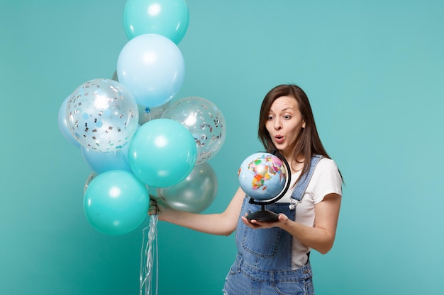 Mujer joven sorprendida con ropa de mezclilla mirando el globo terráqueo en la mano, celebrando con coloridos globos de aire aislados en un fondo azul turquesa. Fiesta de cumpleaños, concepto de emociones de la gente.