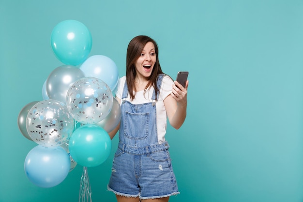 Mujer joven sorprendida con ropa de mezclilla mirando en el teléfono móvil celebrando, sosteniendo coloridos globos de aire aislados en el fondo azul turquesa de la pared. Fiesta de cumpleaños, concepto de emociones de la gente.