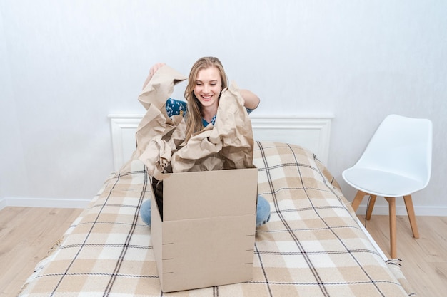 Mujer joven sorprendida sentada en la cama desempacando una caja de cartón en casa Entregando un paquete