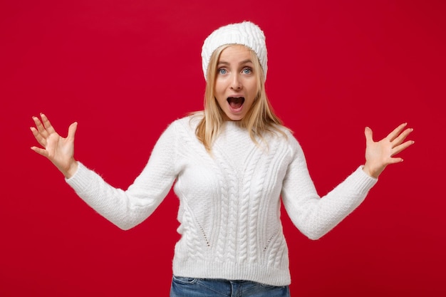 Mujer joven sorprendida con un sombrero de suéter blanco aislado en un fondo rojo en el estudio. Concepto de temporada fría de estilo de vida de moda saludable. Simulacros de espacio de copia. Gesticulando demostrando tamaño con espacio de trabajo horizontal.