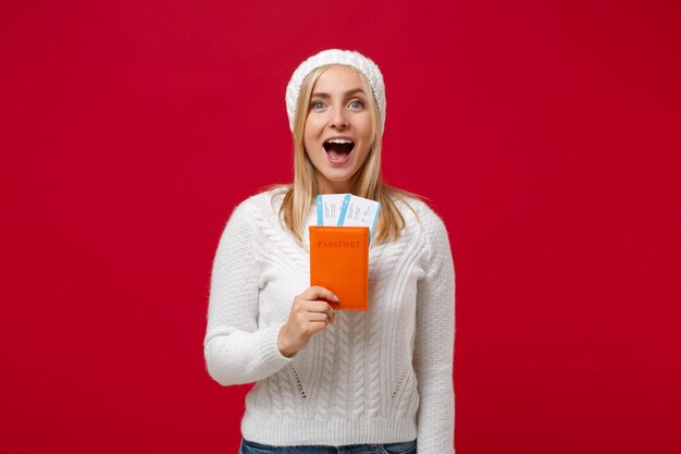 Mujer joven sorprendida con suéter blanco, sombrero aislado en el fondo de la pared roja, retrato de estudio. Estilo de vida de moda saludable, concepto de temporada fría. Simulacros de espacio de copia. Mantenga pasaporte, boletos, tarjeta de embarque.