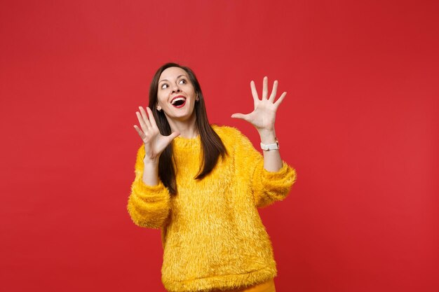 Mujer joven sorprendida en suéter de piel amarilla manteniendo la boca abierta, mirando hacia arriba, extendiendo las manos, dedos aislados sobre fondo de pared roja. Personas sinceras emociones, concepto de estilo de vida. Simulacros de espacio de copia.