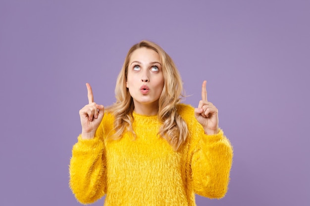 Mujer joven sorprendida con suéter de piel amarilla posando aislada en un retrato de estudio de fondo violeta pastel. Gente emociones sinceras concepto de estilo de vida. Simulacros de espacio de copia. Señalando con los dedos índices hacia arriba.