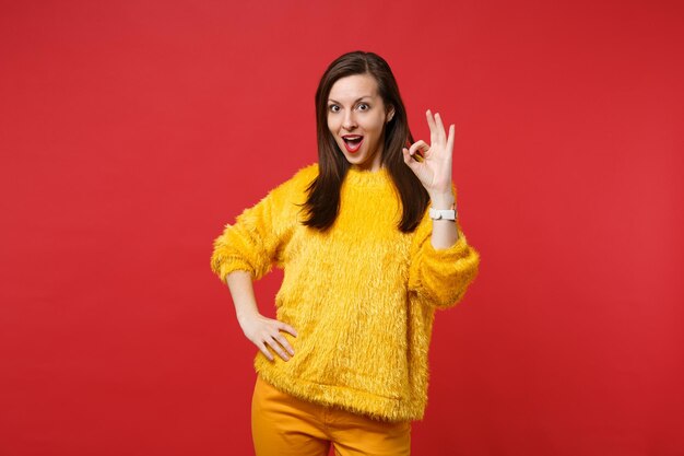 Mujer joven sorprendida en suéter de piel amarillo manteniendo la boca abierta, mostrando gesto OK aislado sobre fondo rojo brillante de la pared en el estudio. Personas sinceras emociones, concepto de estilo de vida. Simulacros de espacio de copia.