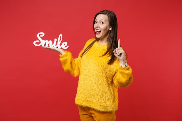 Mujer joven sorprendida en suéter de piel apuntando con el dedo índice hacia arriba con una gran idea nueva mantenga letras de madera palabra sonrisa aislada sobre fondo rojo. Concepto de estilo de vida de emociones sinceras de personas. Simulacros de espacio de copia.