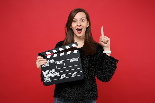 Mujer joven sorprendida en suéter de piel negra manteniendo el dedo índice hacia arriba con una gran idea nueva, sostenga la clásica claqueta de fabricación de película negra aislada sobre fondo rojo. Personas sinceras emociones, concepto de estilo de vida.