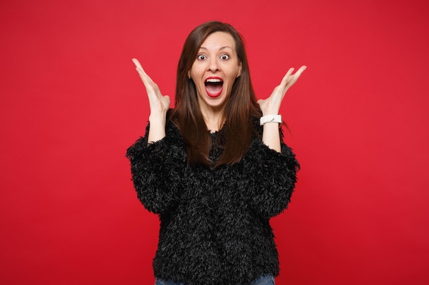 Mujer joven sorprendida en suéter de piel negro manteniendo la boca abierta extendiendo las manos aisladas sobre fondo rojo brillante de la pared, retrato de estudio. Personas sinceras emociones, concepto de estilo de vida. Simulacros de espacio de copia.