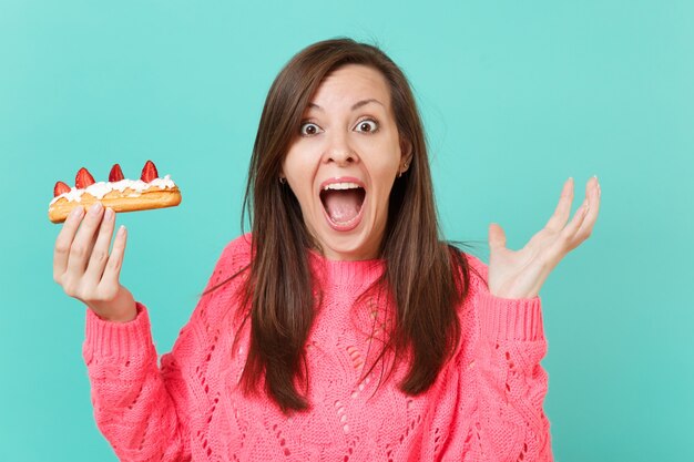 Mujer joven sorprendida en suéter rosa de punto gritando, extendiendo las manos, sosteniendo pastel de eclair aislado sobre fondo de pared azul turquesa, retrato de estudio. Concepto de estilo de vida de personas. Simulacros de espacio de copia.