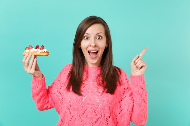Mujer joven sorprendida en suéter rosa tejido manteniendo la boca abierta de par en par, señalando el dedo índice a un lado sostenga en la mano pastel de eclair aislado sobre fondo de pared azul. Concepto de estilo de vida de personas. Simulacros de espacio de copia.