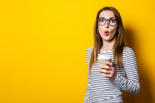 Mujer joven sorprendida en vasos con un café sobre un fondo amarillo