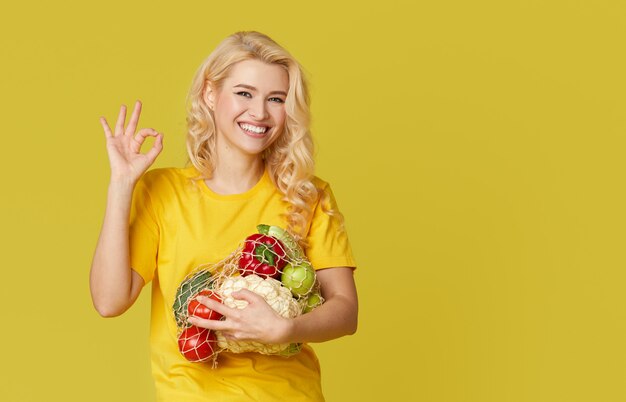 Mujer joven sostenga una bolsa de cadena llena de verduras orgánicas en la pared amarilla.