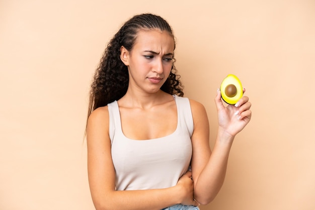 Mujer joven sosteniendo un aguacate aislado sobre fondo beige con expresión triste